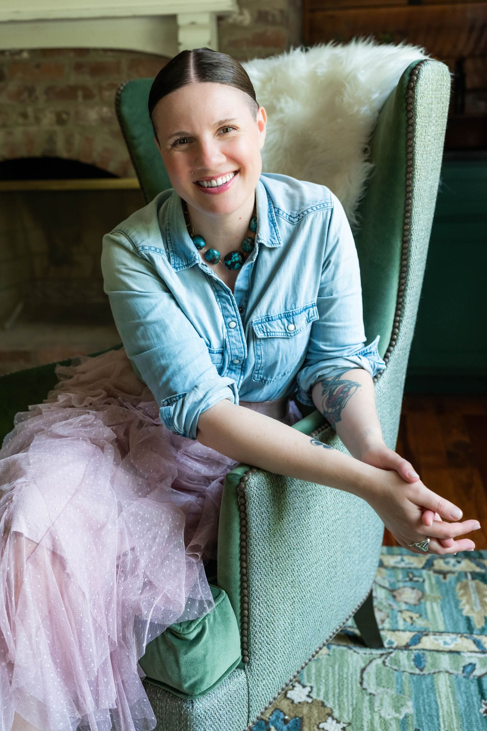 Macon interior designer Lesley Myrick sitting in a custom green wingback chair with nailheads, in a cozy lounge with brick fireplace. She is wearing a chambray shirt and ruffled tulle skirt, along with a bold turquoise and gold necklace.
