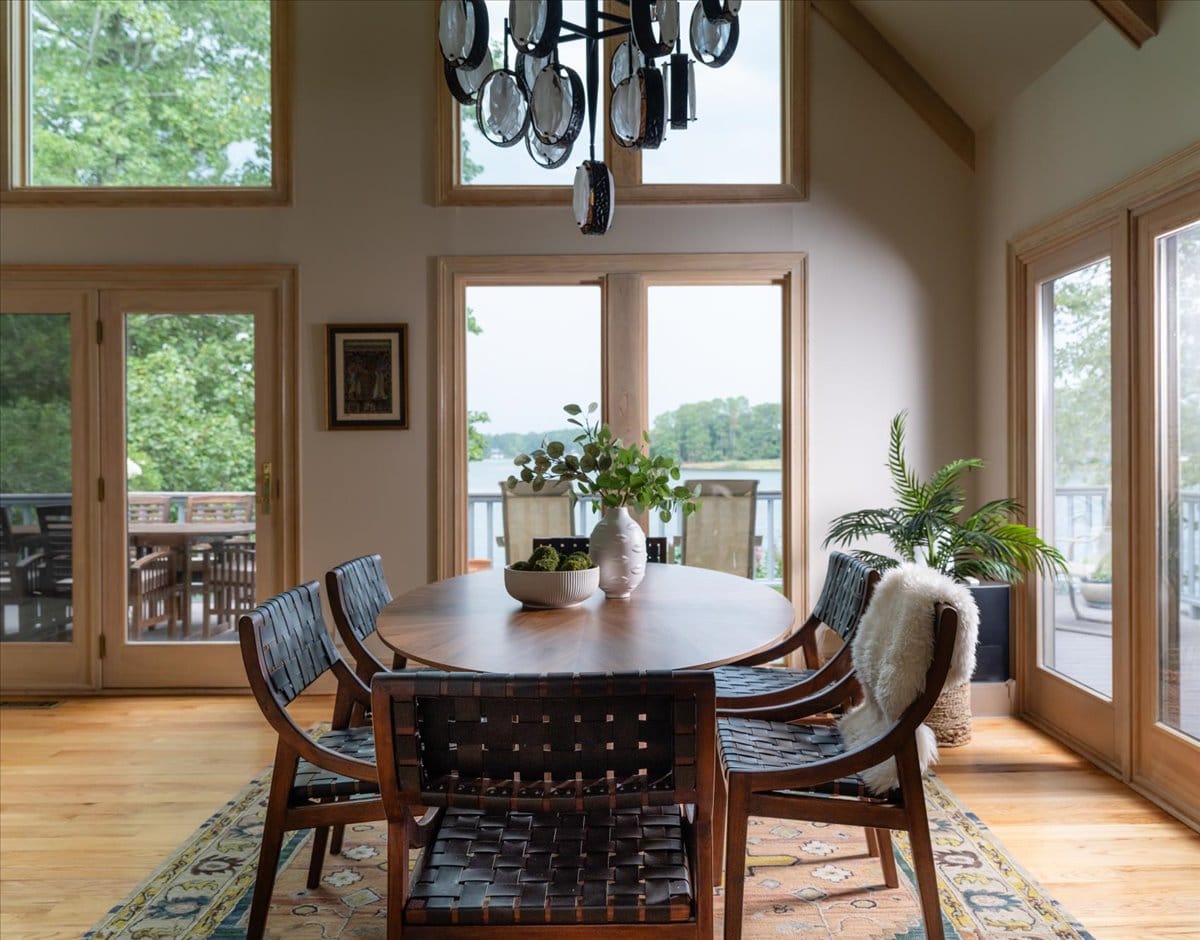 Lakefront dining room with woven leather chairs, oval wood table, modern chandelier, and large windows overlooking the water by Lesley Myrick Interior Design