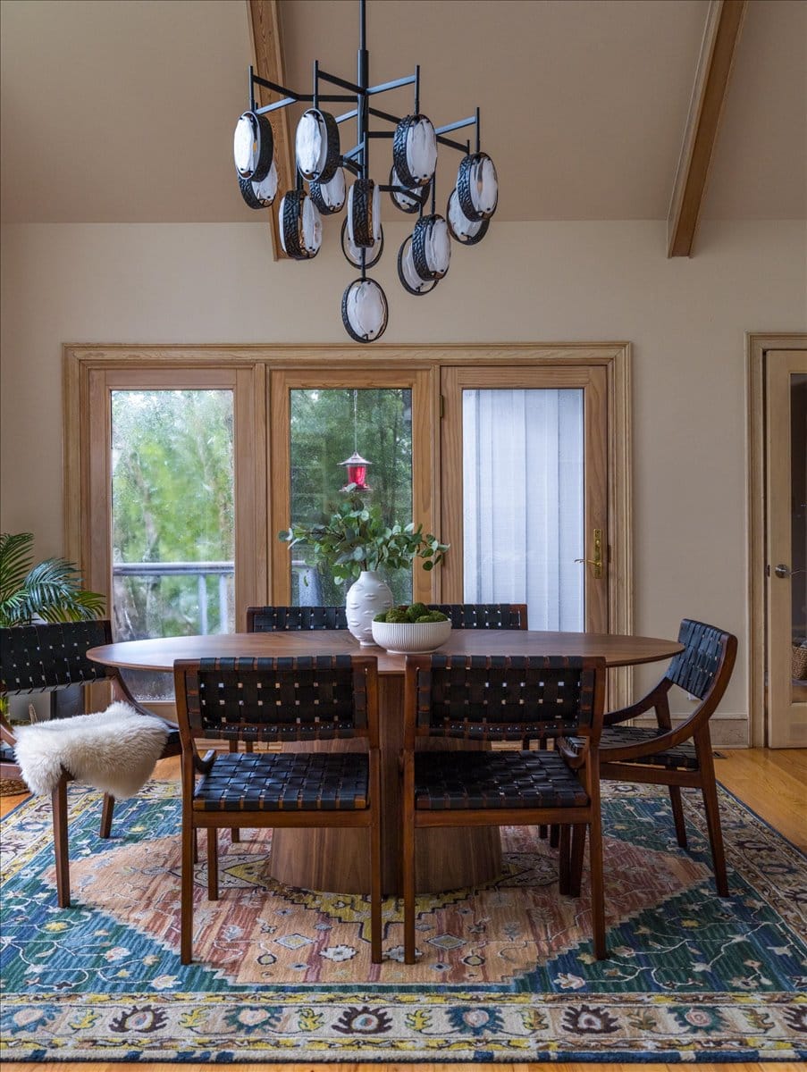 Dining room with oval wood table, black woven leather chairs, colorful patterned rug, and modern chandelier by Lesley Myrick Interior Design