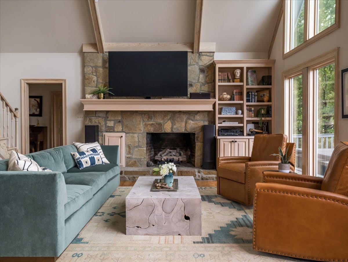Modern rustic living room with teal velvet sofa, tan leather armchairs, stone fireplace, and large windows by Lesley Myrick Interior Design.