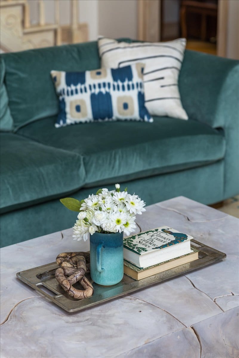 Learn home maintenance tips for a coffee table with stone surface, blue ceramic vase of white flowers, stacked books, and decorative object, with teal velvet sofa in the background by Lesley Myrick Interior Design