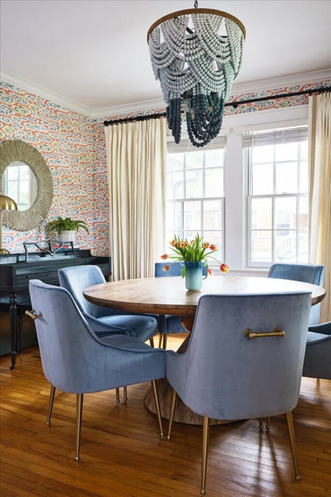 A colorful dining room in a modern Macon bungalow with orange, yellow, and blue cloud print wallpaper, blue velvet dining chairs, and a blue beaded chandelier.