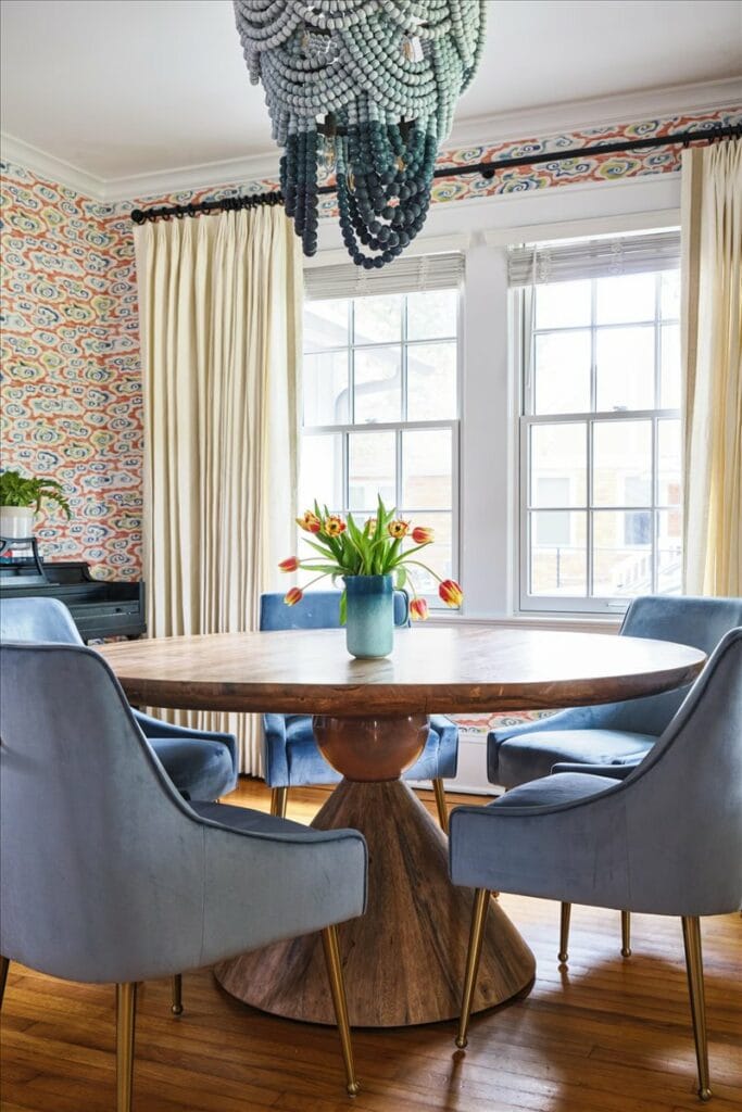 A colorful dining room in a modern Macon bungalow with orange, yellow, and blue cloud print wallpaper, blue velvet dining chairs, and a blue beaded chandelier.