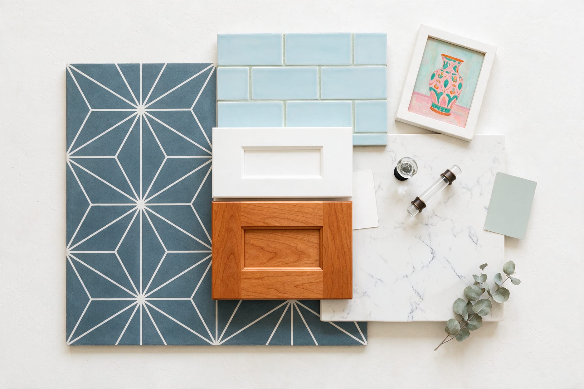 Renovate your home without the stress by selecting all finishes in advance. Overhead flat lay of kitchen renovation material selections including blue geometric floor tile, pale blue subway tile, white and cherry Shaker cabinet doors, marble-look quartz, clear glass hardware, and paint samples